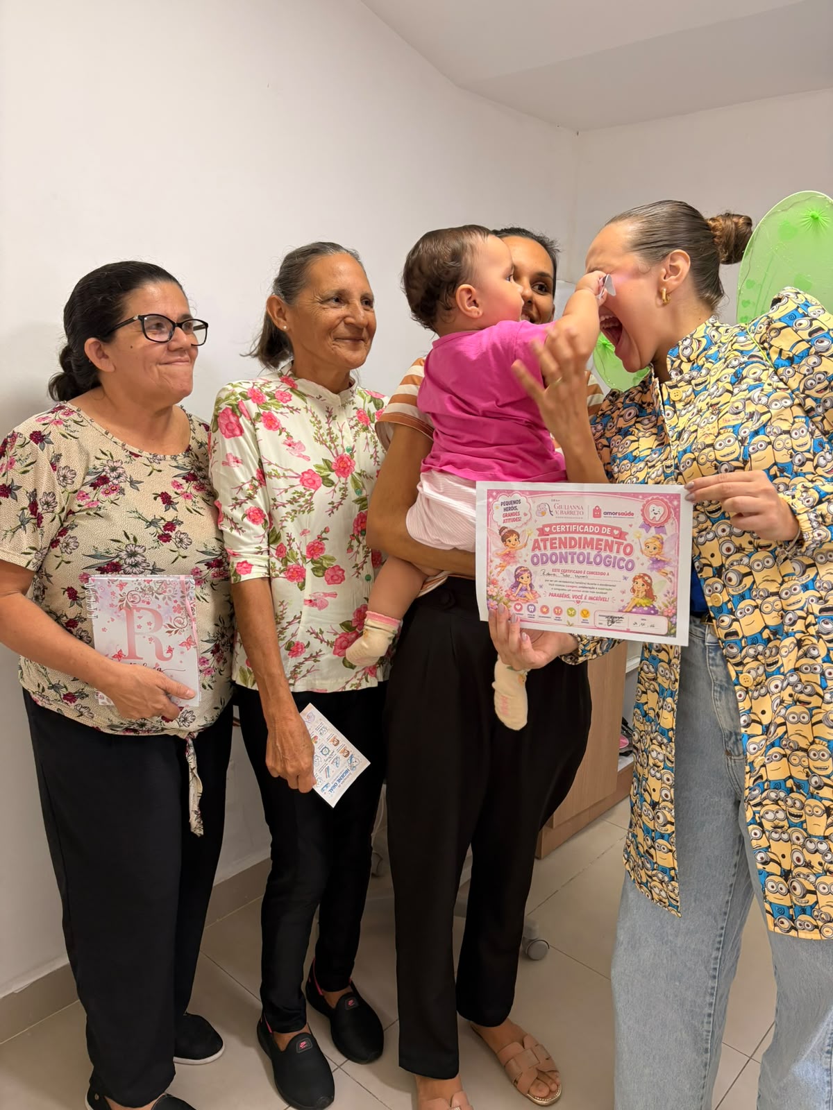 Dra. Giulianna Barreto com um bebê no colo da mãe, no consultório, ao lado do certificado de atendimento odontológico