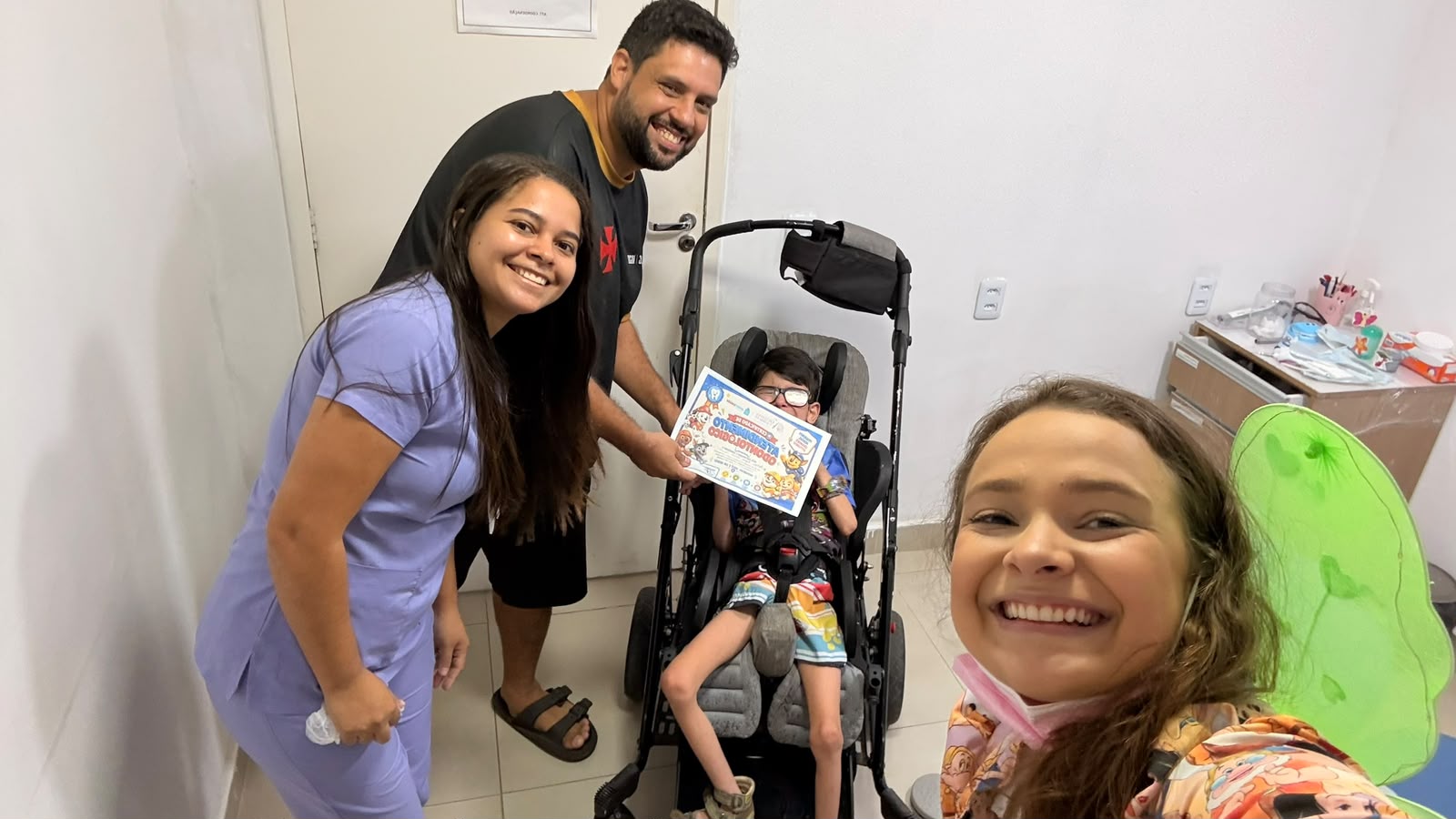 Dra. Giulianna Barreto, com asas de fada, ao lado de uma família e de uma criança em cadeira de rodas adaptada