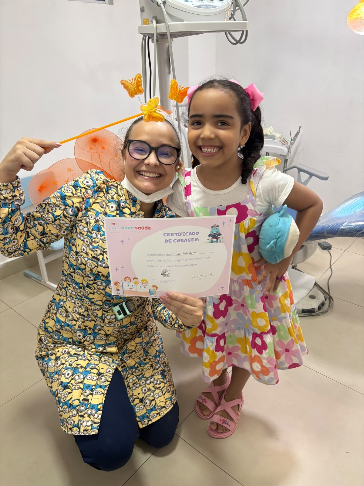 Menina sorrindo ao lado da Dra. Giulianna Barreto, segurando seu Certificado de Coragem