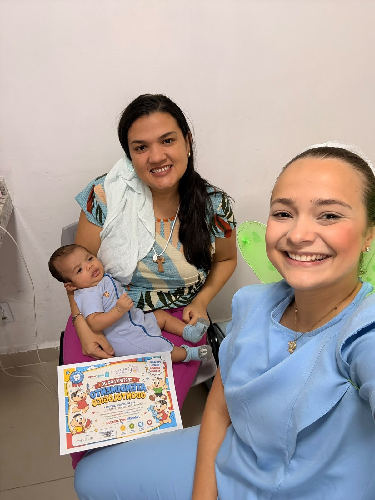 Dra. Giulianna Barreto com uma mãe e seu bebê, segurando o certificado de atendimento odontológico
